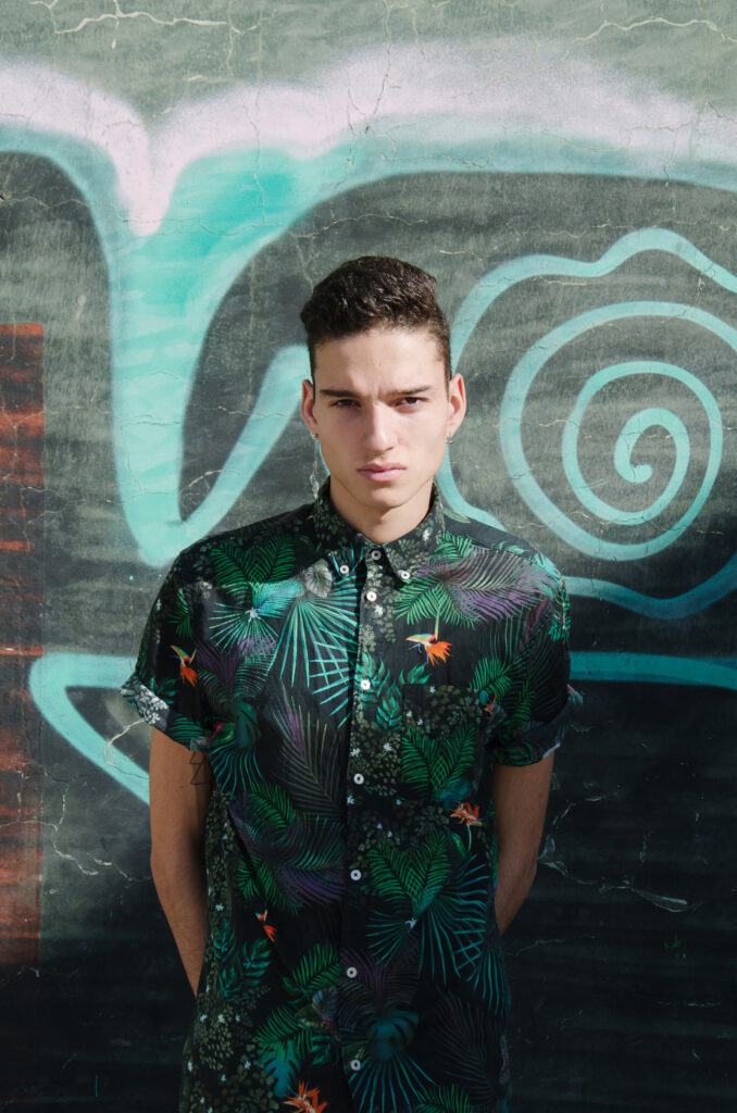 Victor Zepeda stands against a graffiti-covered wall wearing a vibrant tropical-patterned shirt with a focused and intense expression on his face.